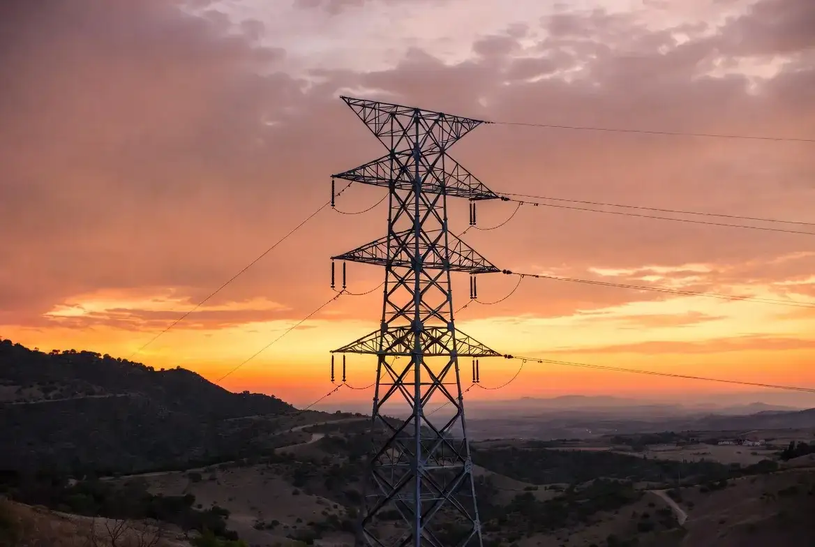 Torre de alta tensión recortada contra un cielo de atardecer naranja sobre paisaje montañoso